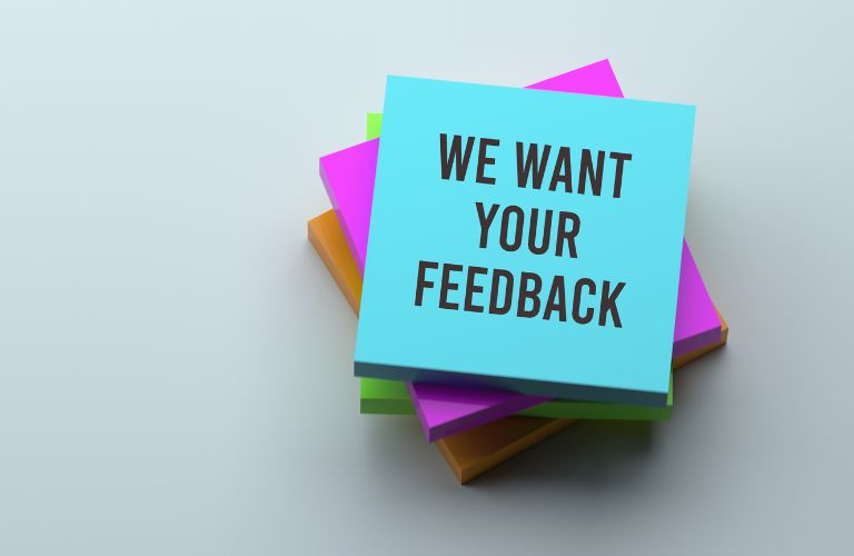 We Want Your Feedback Text on Blue Post-It Note in a Pile of Post-It Notes